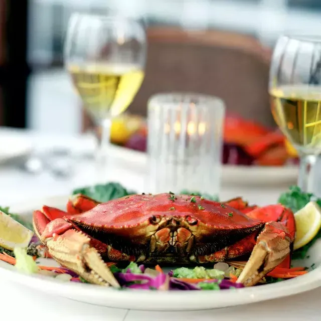 A Dungeness crab sits on a plate in a 餐厅 with two glasses of white wine 在 background.